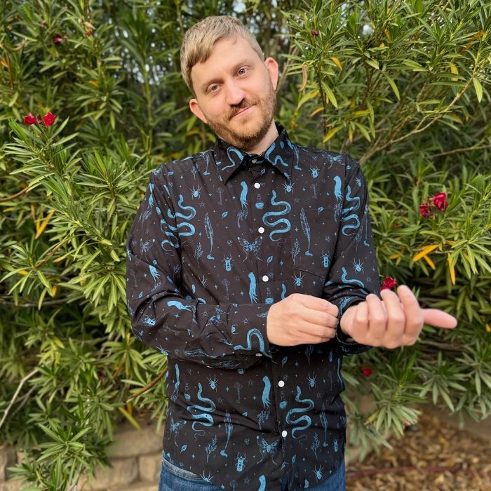 Man in a dark patterned button up shirt standing in front of green shrubs