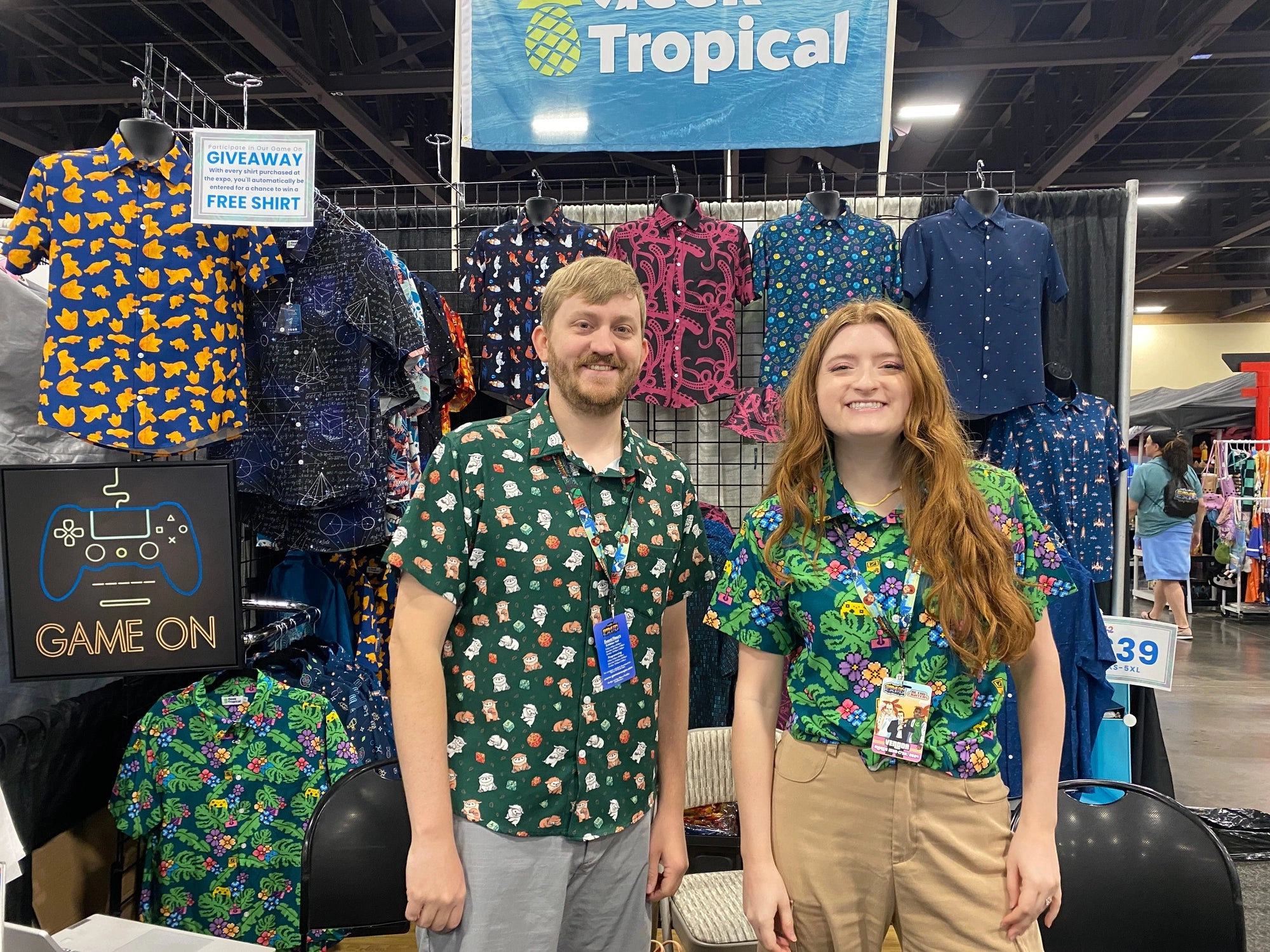 Two Geek Tropical team members in matching tropical-print shirts at a convention booth, with a wall of button up shirts behind them.